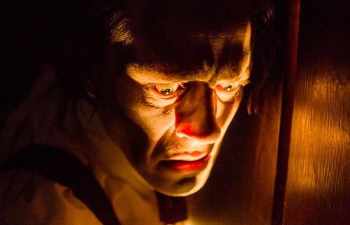 A man's ghoulish face is lit by an orange glow, coming from beneath the floorboards.